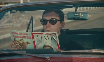 Movie still from “A Man and a Woman” (1966), directed by Claude Lelouch – A man sitting in the back seat of a car reading a magazine; Medium shot, Low angle