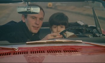 Movie still from “A Man and a Woman” (1966), directed by Claude Lelouch – A man and a boy in a car looking at a book; Medium shot, High angle