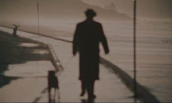 Movie still from “A Man and a Woman” (1966), directed by Claude Lelouch – A man in a long coat and a hat walking a dog on a leash; Wide shot, Low angle