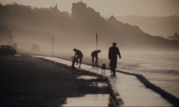 Movie still from “A Man and a Woman” (1966), directed by Claude Lelouch – Three people are walking along the sidewalk near the water; Extreme Wide shot, Low angle