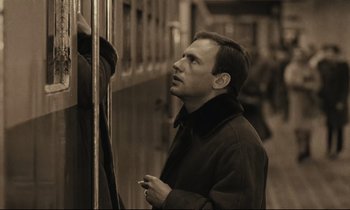 Movie still from “A Man and a Woman” (1966), directed by Claude Lelouch – A black and white photo of a man standing next to a fence; Close Up shot, Over the shoulder angle