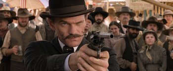 Movie still from “A Million Ways to Die in the West” (2014), directed by Seth MacFarlane – A man in a hat and a suit is holding a gun; Close Up shot, Over the shoulder angle