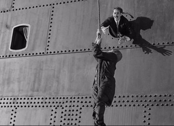 Movie still from “A Night at the Opera” (1935), directed by Sam Wood – A man hanging from a metal wall while another man holds onto a rope; Wide shot, Overhead angle