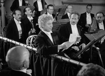 Movie still from “A Night at the Opera” (1935), directed by Sam Wood – An old photo of a man holding a sheet of paper in front of an orchestra; Medium shot, Low angle