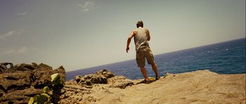 Movie still from “A Perfect Getaway” (2009), directed by David Twohy – A man standing on top of a rock near the ocean; Wide shot, Low angle