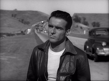 Movie still from “A Place in the Sun” (1951), directed by George Stevens – A black and white photo of a man standing on the side of a road; Medium shot, Over the shoulder angle