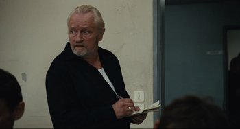 Movie still from “A Prophet” (2009), directed by Jacques Audiard – An older man holding a book while standing next to a wall; Medium shot, Over the shoulder angle