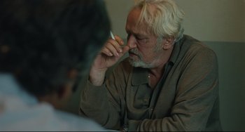 Movie still from “A Prophet” (2009), directed by Jacques Audiard – An older man with white hair and a gray beard is holding a pen to his nose; Close Up shot, Over the shoulder angle