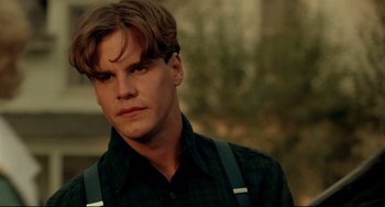 Movie still from “A River Runs Through It” (1992), directed by Robert Redford – A young man with long brown hair wearing a black shirt and suspenders; Close Up shot, Over the shoulder angle