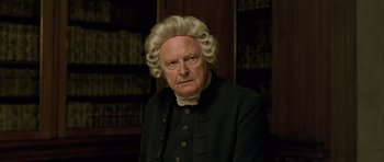 Movie still from “A Royal Affair” (2012), directed by Nikolaj Arcel – An old man with a wig and a beard in front of a book case; Close Up shot, Over the shoulder angle