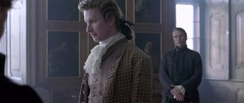 Movie still from “A Royal Affair” (2012), directed by Nikolaj Arcel – A man in a brown and white suit standing in front of a wall; Medium shot, Over the shoulder angle