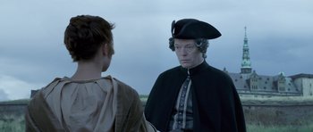 Movie still from “A Royal Affair” (2012), directed by Nikolaj Arcel – A man wearing a hat standing next to a woman; Medium shot, Over the shoulder angle