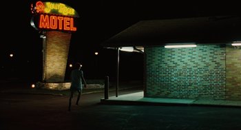 Movie still from “A Serious Man” (2009), directed by Joel Coen – A person walking on the sidewalk near a motel sign at night; Wide shot, Over the shoulder angle