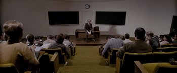 Movie still from “A Single Man” (2009), directed by Tom Ford – A man sitting at a desk in front of a group of people; Wide shot, High angle