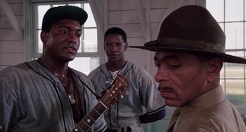 Movie still from “A Soldier's Story” (1984), directed by Norman Jewison – A group of men standing next to each other with a guitar; Medium shot, Over the shoulder angle