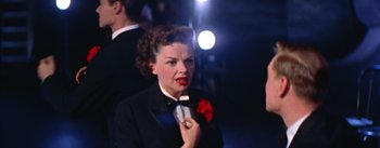 Movie still from “A Star Is Born” (1954), directed by George Cukor – A woman in a black suit and a red flower in her lapel; Close Up shot, Low angle