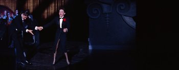 Movie still from “A Star Is Born” (1954), directed by George Cukor – A woman in a black suit and a red flower in her hand; Wide shot, Low angle