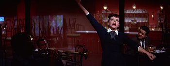 Movie still from “A Star Is Born” (1954), directed by George Cukor – A woman in a black dress is holding her arms up; Medium shot, Low angle