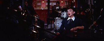 Movie still from “A Star Is Born” (1954), directed by George Cukor – A woman in a suit and tie singing in front of a band; Medium shot, High angle