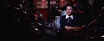 Movie still from “A Star Is Born” (1954), directed by George Cukor – A woman sitting at a table in front of a microphone; Medium shot, Over the shoulder angle
