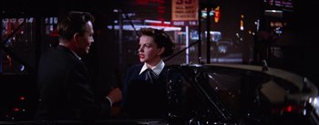 Movie still from “A Star Is Born” (1954), directed by George Cukor – A woman standing next to a car on a city street at night; Medium shot, Over the shoulder angle