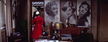 Movie still from “A Star Is Born” (1954), directed by George Cukor – A woman in a red dress standing in front of two black and white pictures; Medium shot, Low angle