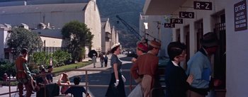 Movie still from “A Star Is Born” (1954), directed by George Cukor – A group of people walking down a street near a building; Wide shot, Over the shoulder angle