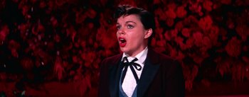 Movie still from “A Star Is Born” (1954), directed by George Cukor – A woman in a suit and bow tie is singing; Close Up shot, Low angle