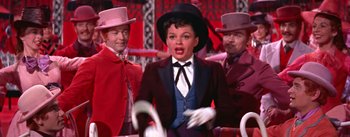 Movie still from “A Star Is Born” (1954), directed by George Cukor – A group of men standing next to a woman in a suit; Medium shot, High angle