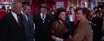 Movie still from “A Star Is Born” (1954), directed by George Cukor – An older man and a woman are standing in a crowd; Medium shot, Over the shoulder angle
