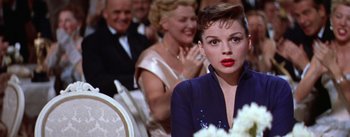 Movie still from “A Star Is Born” (1954), directed by George Cukor – A woman sitting in front of a group of other people; Close Up shot, Over the shoulder angle