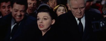 Movie still from “A Star Is Born” (1954), directed by George Cukor – A group of people that are standing in a room; Close Up shot, Low angle