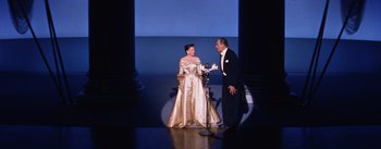 Movie still from “A Star Is Born” (1954), directed by George Cukor – A man and a woman in formal wear standing in front of a microphone; Wide shot, High angle