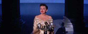 Movie still from “A Star Is Born” (1954), directed by George Cukor – A woman in a gold dress is holding a microphone; Medium shot, Low angle