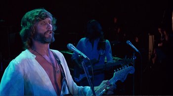 Movie still from “A Star Is Born” (1976), directed by Frank Pierson – A man holding a microphone while playing a guitar in front of other men; Medium shot, Low angle