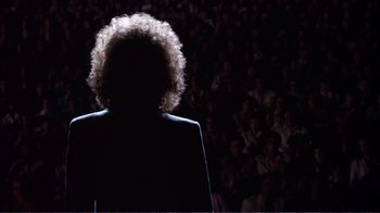 Movie still from “A Star Is Born” (1976), directed by Frank Pierson – A person with curly hair standing in front of an audience; Wide shot, Over the shoulder angle