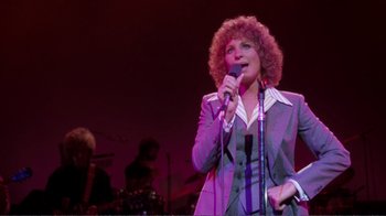 Movie still from “A Star Is Born” (1976), directed by Frank Pierson – A woman with curly hair is singing into a microphone on a stage; Medium shot, High angle