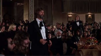 Movie still from “A Star Is Born” (1976), directed by Frank Pierson – A group of people sitting in a room with a man in a tuxedo holding a cell phone; Wide shot, Over the shoulder angle