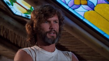 Movie still from “A Star Is Born” (1976), directed by Frank Pierson – A man with long hair and a beard is standing in front of a stained glass window; Close Up shot, Low angle