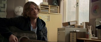 Movie still from “A Street Cat Named Bob” (2016), directed by Roger Spottiswoode – A man sitting in front of a guitar in a room; Medium shot, Over the shoulder angle