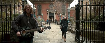 Movie still from “A Street Cat Named Bob” (2016), directed by Roger Spottiswoode – A woman walking down a street while holding a guitar; Wide shot, Over the shoulder angle