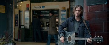 Movie still from “A Street Cat Named Bob” (2016), directed by Roger Spottiswoode – A man playing a guitar in front of a ticket booth; Medium shot, Over the shoulder angle