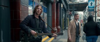 Movie still from “A Street Cat Named Bob” (2016), directed by Roger Spottiswoode – A man holding a guitar on the side of the street; Medium shot, Over the shoulder angle