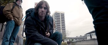 Movie still from “A Street Cat Named Bob” (2016), directed by Roger Spottiswoode – A man sitting on the side of the road with a backpack; Medium shot, Low angle