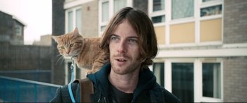 Movie still from “A Street Cat Named Bob” (2016), directed by Roger Spottiswoode – A man with long brown hair and blue eyes is holding a cat on his shoulder; Close Up shot, Over the shoulder angle