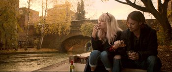 Movie still from “A Street Cat Named Bob” (2016), directed by Roger Spottiswoode – A woman smoking a cigarette while sitting next to a river; Medium shot, High angle