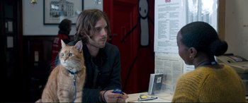 Movie still from “A Street Cat Named Bob” (2016), directed by Roger Spottiswoode – A man sitting at a table with a cat on top of it; Medium shot, Over the shoulder angle