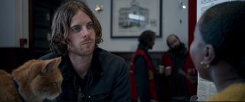 Movie still from “A Street Cat Named Bob” (2016), directed by Roger Spottiswoode – A man with long hair sitting at a table with two other men; Close Up shot, Over the shoulder angle