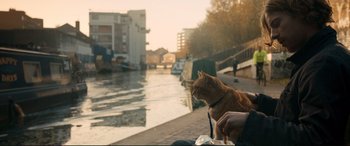 Movie still from “A Street Cat Named Bob” (2016), directed by Roger Spottiswoode – An orange cat sitting on a person's lap next to a river; Medium shot, Over the shoulder angle