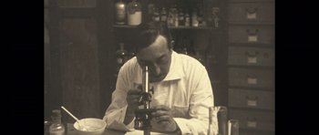Movie still from “A Very Long Engagement” (2004), directed by Jean-Pierre Jeunet – An old black and white photo of a man using a microscope; Medium shot, High angle